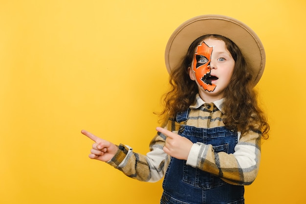 El promotor sorprendió a la niña pequeña con máscara de maquillaje de Halloween que usa sombrero y camisa, señala con el dedo a un lado en el área de promoción de la maqueta del espacio de copia del espacio de trabajo, posando aislado sobre fondo de color amarillo en el estudio