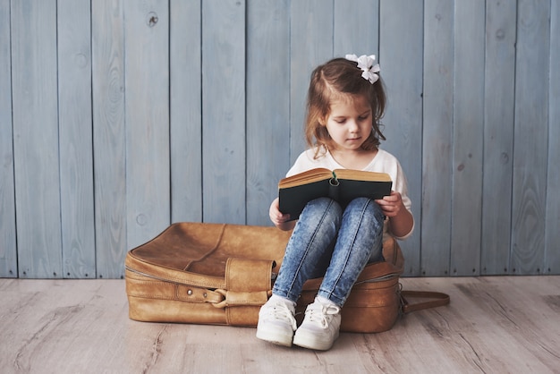 Pronto para grandes viagens. Menina feliz lendo livro interessante, carregando uma maleta grande e sorrindo. Viagem, liberdade e imaginação