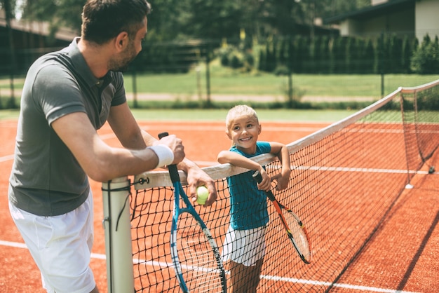 Pronto para jogar? Pai e filha alegres inclinando-se para a rede de tênis e olhando um para o outro com sorrisos