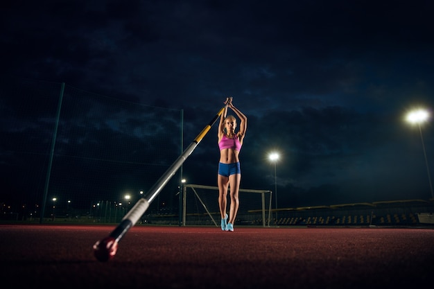 Pronto para superar as dificuldades. Treinamento profissional de salto com vara feminino no estádio à noite. Praticar ao ar livre. Conceito de esporte