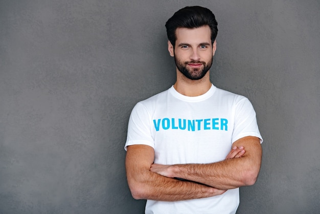 Pronto para trabalhos de caridade. Jovem confiante com uma camiseta de voluntário, mantendo os braços cruzados e olhando para a câmera com um sorriso em pé contra um fundo cinza