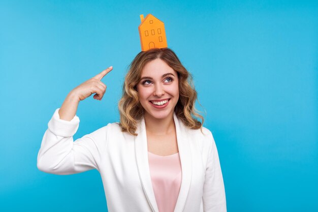 Propiedad de la casa. Retrato de mujer alegre con cabello ondulado en chaqueta blanca apuntando a una pequeña casa de papel en la cabeza y sonriendo ampliamente, soñando con su propio apartamento. foto de estudio interior, fondo azul