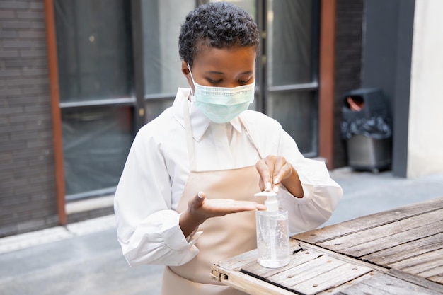 La propietaria de una pequeña empresa africana negra hace un trabajo de preparación para la reapertura del negocio usando una máscara facial adecuada y limpiándose la mano con un desinfectante de manos con gel de alcohol antes de comenzar a trabajar