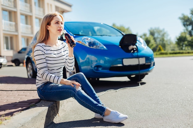 Propietario de auto. Mujer joven bastante encantada sentada frente a su coche y bebiendo refrescos mientras lo espera en la estación de carga