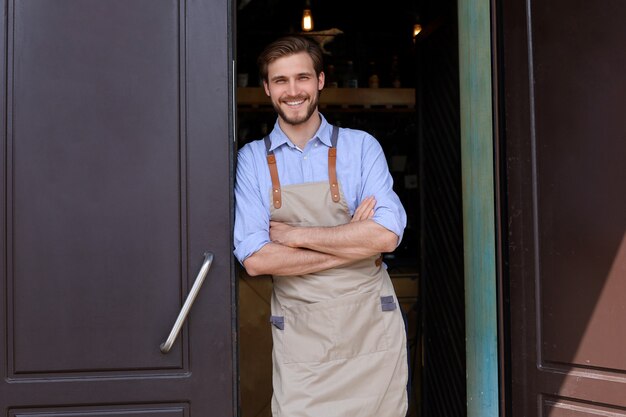 Propietario de café guapo y seguro de pie en la puerta. Hombre joven de pie con los brazos cruzados mirando a la cámara sonriendo.