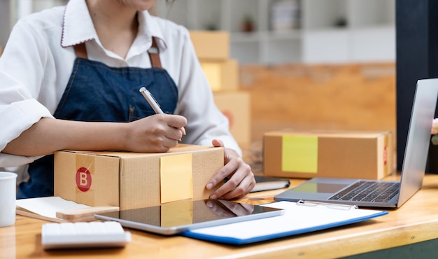 Propietario de envío de compras en línea escribiendo la dirección en una caja de cartón en el lugar de trabajo empresario de pequeñas empresas PYME o mujer asiática independiente que trabaja con una caja en casa