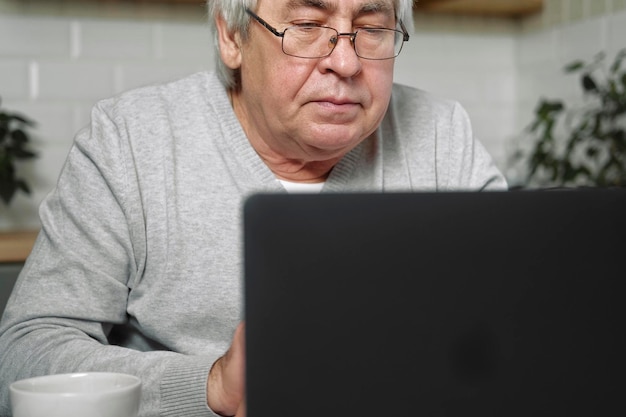 Propietario de un negocio masculino mayor con anteojos sentado en el escritorio frente a la computadora portátil Hombre mayor exitoso trabajando en casa Cabello gris 60s 70s Anciano ocupado escribiendo teclado bebiendo café Oficina a distancia