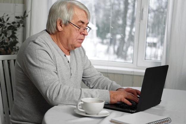 Propietario de un negocio masculino mayor con anteojos sentado en el escritorio frente a la computadora portátil Hombre mayor exitoso trabajando en casa Cabello gris 60s 70s Anciano ocupado escribiendo teclado mirando la pantalla Oficina a distancia