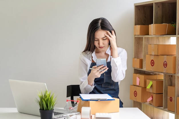 Propietario de negocio de mujer asiática sosteniendo un teléfono inteligente estrés dolores de cabeza con ordenar con caja de paquetería en casa.