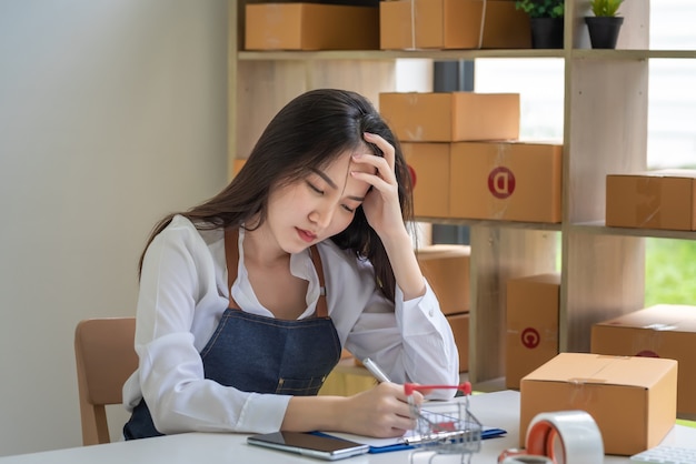 Propietario de una pequeña empresa en línea Mujer asiática con delantal hizo hincapié en el dolor de cabeza tomando notas con la caja de paquetería en casa.