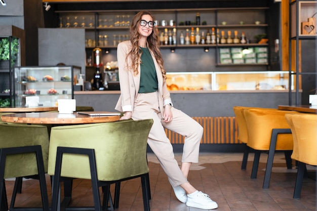 Propietario de restaurante de mujer de negocios vestido con traje de pantalón elegante de pie en el restaurante con fondo de barra de bar Gafas de mujer caucásica Persona de negocios Manos interiores en bolsillos de longitud completa