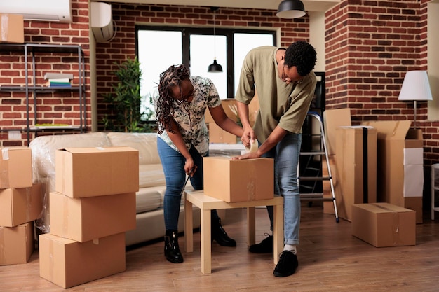 Propietarios casados que preparan muebles para transportarlos a un nuevo apartamento alquilado, usando rodillos de cinta adhesiva para empacar cosas en cajas. Mudanza en casa familiar comprada con préstamo hipotecario.