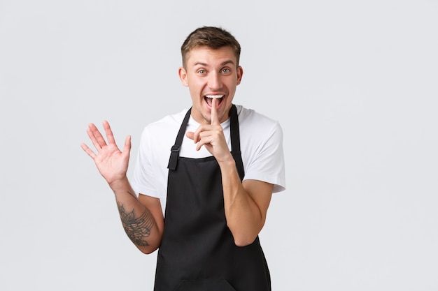 Propietarios de pequeñas empresas, cafetería y concepto de personal. Guapo barista feliz, camarero en la cafetería con delantal negro, riendo y haciendo un gesto de silencio, pidiendo silencio o no decir en secreto