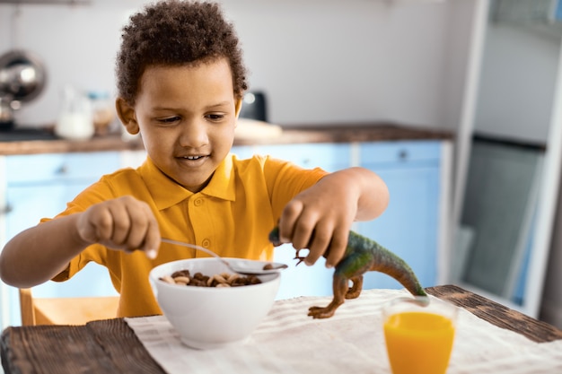 Proprietário atencioso. Garotinho otimista sentado à mesa da cozinha tomando café da manhã enquanto dá cereais para seu dinossauro de brinquedo