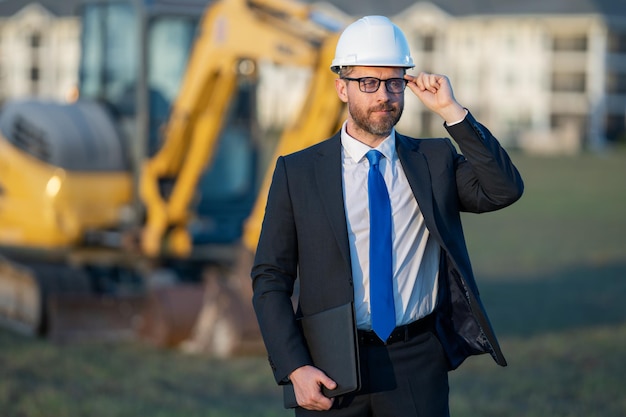 Proprietário de construção bem-sucedido, trabalhador da construção civil de terno e capacete perto da escavadeira confidencia