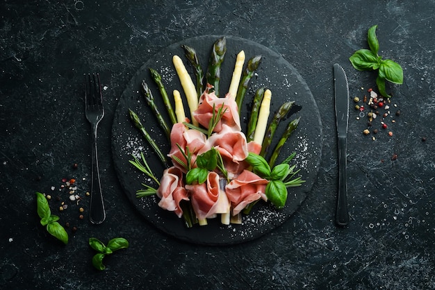 Prosciutto mit gekochtem Spargel und Gewürzen auf einer schwarzen Steinplatte Draufsicht Freier Platz für Ihren Text