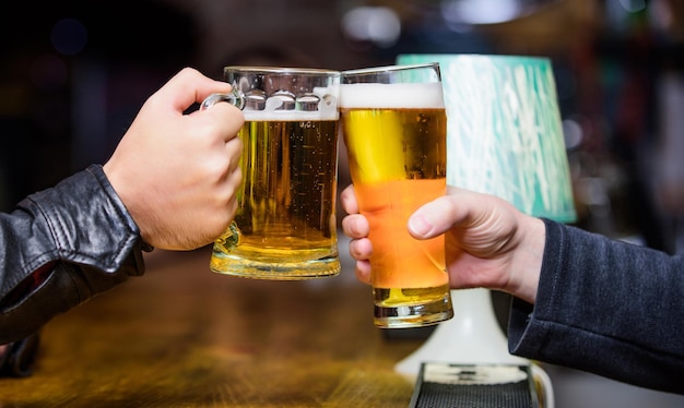 Prost-Konzept. Glas mit frischem Lagerbier vom Fass mit Schaum. Männliche Hände halten Tassen mit kaltem, leckerem Bier in der Bar. Freitag Freizeittradition. Bierkrüge an der Theke defokussierten Hintergrund.