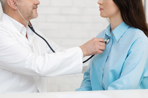 Proteção da saúde, exame do paciente e visita ao médico de família. Médico adulto e amigável, de jaleco branco, ouve com estetoscópio mulheres no consultório, vista lateral, panorama, espaço livre, cortado