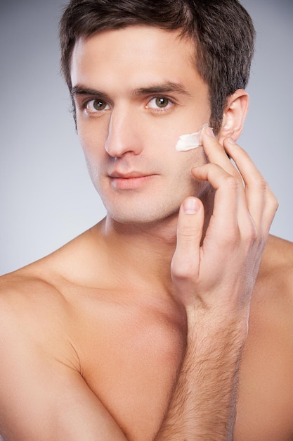 Protección de la piel. Apuesto joven sin camisa aplicando crema en la cara y mirando a la cámara mientras está de pie aislado sobre fondo gris