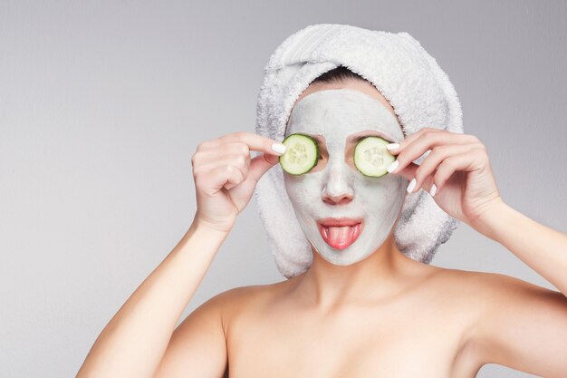 Protección de la piel. Hermosa chica con máscara de colágeno y rodajas de pepino en los ojos divirtiéndose contra el fondo gris, obteniendo un tratamiento de belleza. Concepto de spa con espacio de copia