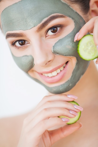 Protección de la piel. Mujer joven con máscara de arcilla cosmética con pepino en su baño