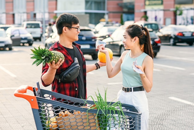 Provável casal asiático em pé perto do carrinho de compras, homem detém abacaxi e melão e mulher bonita conta as notas de dólares dos EUA.