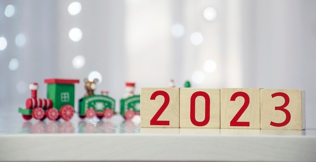 Próximamente Número de Año Nuevo 2023 en bloque de cubo de madera con colorido tren de Navidad y luces bokeh borrosas en el fondo Tarjeta de felicitación de Feliz Navidad y Feliz Año Nuevo para las vacaciones de invierno