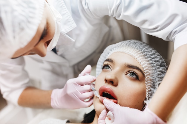 Prozess der Lippenverbesserung. Kosmetikerin macht eine Injektion von Hyaluronsäure in eine schöne Lippen. Das junge Mädchen mit einem schönen Gesicht im speziellen Hut und den Händen des Arztes in den rosa Handschuhen.