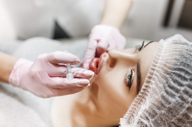 Prozess der Lippenverbesserung. Kosmetikerin macht eine Injektion von Hyaluronsäure in eine schöne Lippen. Das junge Mädchen mit einem schönen Gesicht im speziellen Hut und den Händen des Arztes in den rosa Handschuhen.