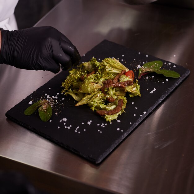 Prozess des Kochens von warmem Salat mit Kalbfleisch. Hände eines Kochs in schwarzen Handschuhen. Schwarzes Schieferbrett.