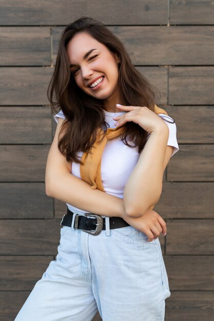 Ptoto vertical de joven hermosa mujer morena feliz positiva de pie contra la pared marrón