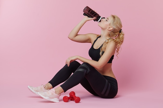 Publicidad entrenamiento personal. Una joven deportiva bebe un batido de proteínas de una coctelera después de entrenar en el interior. pared rosa