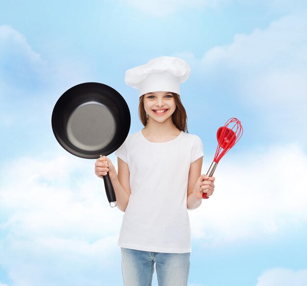 publicidad, infancia, cocina y gente - chica sonriente con camiseta blanca y gorro de cocina sosteniendo una sartén sobre el fondo del cielo nublado