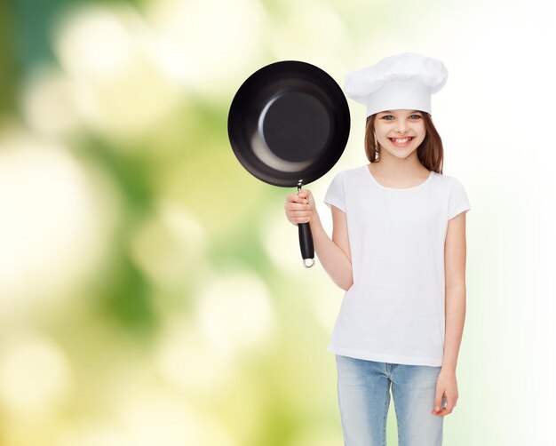 publicidad, infancia, cocina y gente - chica sonriente con camiseta blanca y sombrero de cocina sosteniendo una sartén sobre fondo verde