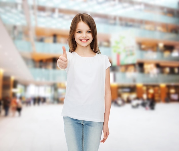 publicidad, infancia, gesto, consumismo y gente - chica sonriente con camiseta blanca mostrando los pulgares hacia arriba sobre el fondo del centro comercial