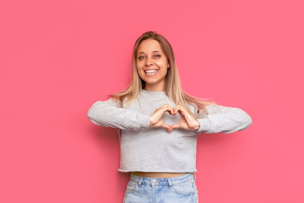 para publicidad Una joven rubia caucásica bastante encantadora formando una forma de corazón con sus manos aisladas en una pared de color rosa brillante Copia espacio para texto o diseño Concepto de caridad