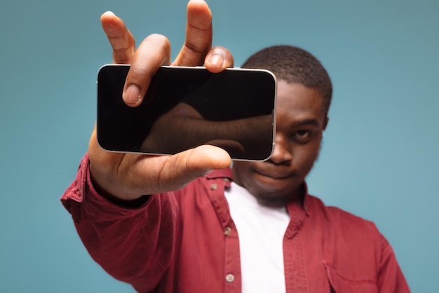 Publicidad móvil Hombre negro muy alegre apuntando a un teléfono celular con una pantalla blanca vacía en una maqueta de fondo de estudio azul para aplicación o sitio web Plantilla de visualización de teléfono celular