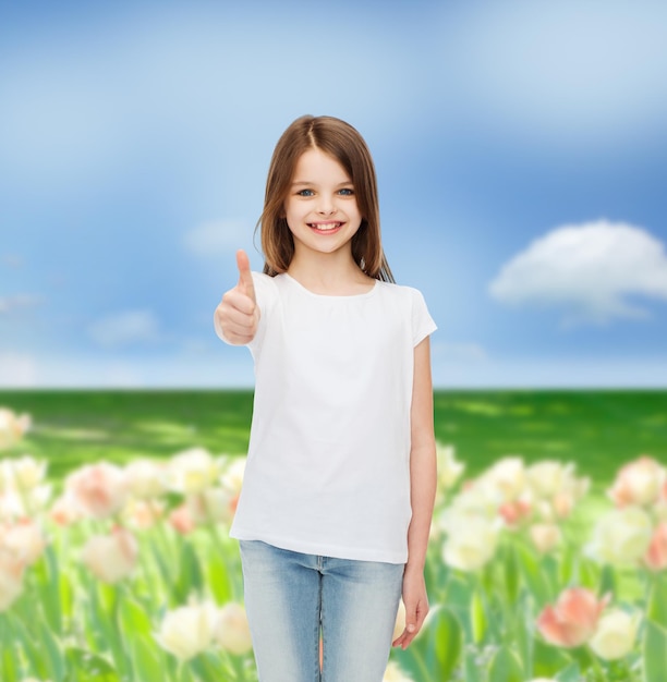 publicidad, niñez, naturaleza, gesto y concepto de la gente - chica sonriente con camiseta blanca mostrando los pulgares hacia arriba sobre el fondo del campo de flores