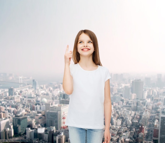 publicidade, infância, gesto e conceito de pessoas - menina sorridente em camiseta branca em branco apontando o dedo para cima sobre o fundo da cidade
