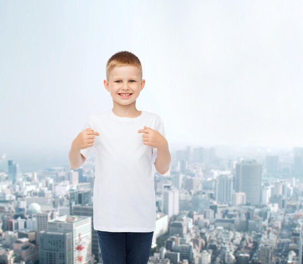 publicidade, pessoas e conceito de infância - menino sorridente em camiseta branca em branco apontando os dedos para si mesmo sobre o fundo da cidade