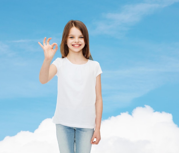 publicidade, sonho, gesto, infância e pessoas - menina sorridente em camiseta branca em branco mostrando sinal de ok sobre fundo de céu azul