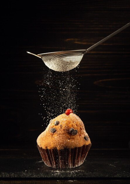 Puderzucker wird aus einem Sieb auf einen gebackenen köstlichen Cupcake gegossen. Das Konzept eines köstlichen Desserts zum Mittagessen auf schwarzem Hintergrund