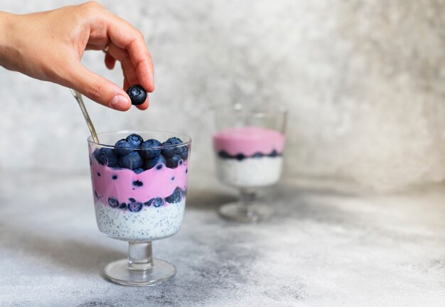 Foto pudim de chia saudável café da manhã com mirtilos, iogurte grego e iogurte de mirtilo natural. feminino mão segurando o pudim de chia. comida saudável, conceito vegetariano. fundo cinza claro