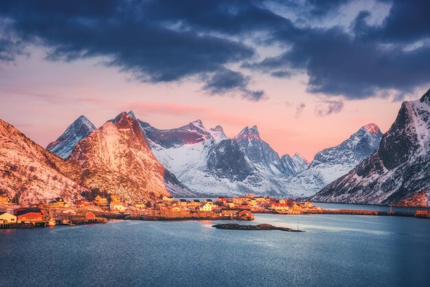 Pueblo de Reine y montañas cubiertas de nieve en el hermoso amanecer en invierno Islas Lofoten Noruega Paisaje con casas de rocas nevadas de mar azul y cielo colorido rorbus tradicional con nubes Costa