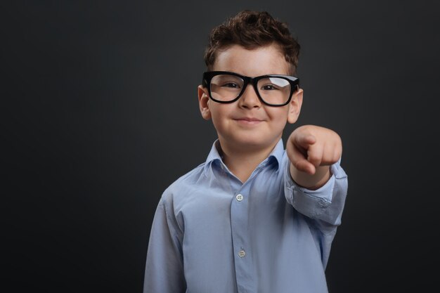 Puedes hacerlo. Dulce chico agradable confiado que te anima a hacer algo mientras usas gafas y se encuentran aisladas sobre fondo gris