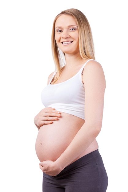 Puedo sentir a mi bebé moverse. Vista lateral de la hermosa mujer embarazada tomados de la mano sobre su abdomen y sonriendo mientras está de pie aislado en blanco