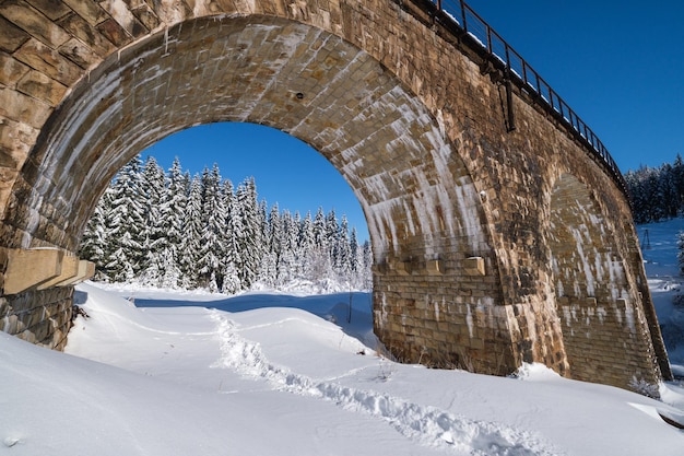 Puente de arco de viaducto de piedra en el ferrocarril a través del bosque de abetos nevados de montaña Ventisqueros en el borde del camino y escarcha en árboles y cables eléctricos