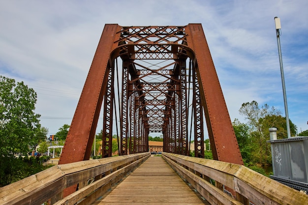 Puente peatonal de hierro marrón con vigas de metal cruzadas bajo un cielo azul nublado