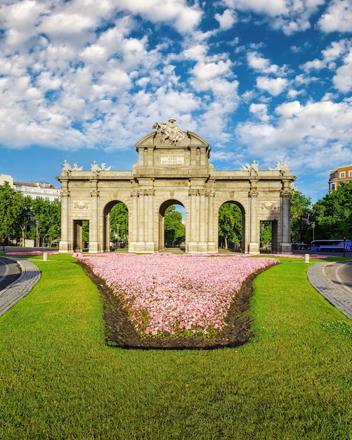 Puerta de Alcalá de Madrid en flores en el suelo y día soleado con nubes blancas.
