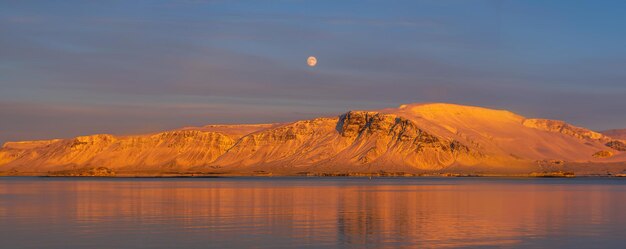 Puesta de sol desde el paseo marítimo de Reykjavik que muestra la luna llena y las cumbres nevadas y anaranjadas